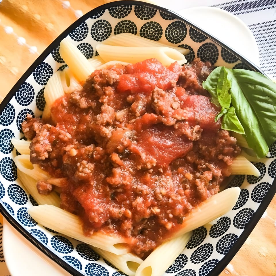 Penne pasta with a rich red meat sauce and fresh basil in a patterned bowl.