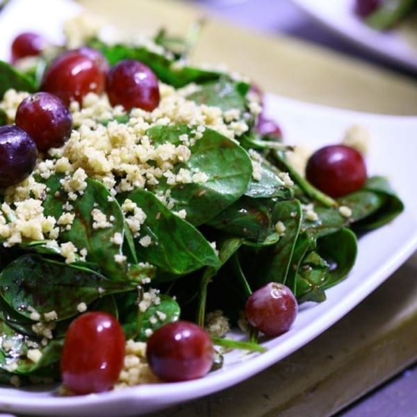 A vibrant spinach salad with red grapes and crumbled cheese on a white plate.