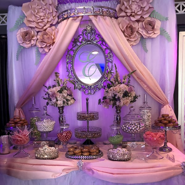 A dessert table with donuts, candies, and lollipops, decorated with pink drapes, flowers, and a mirror.