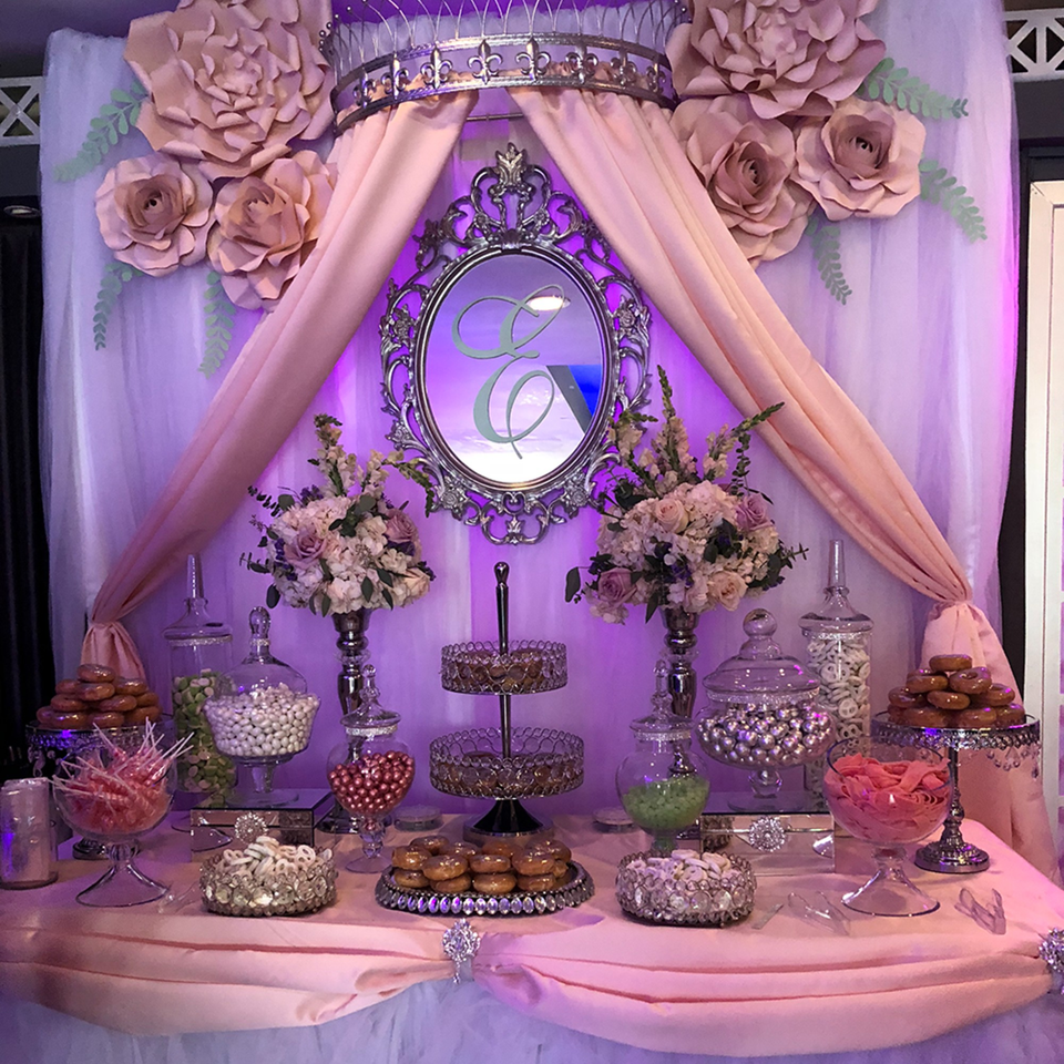 A dessert table with donuts, candies, and lollipops, decorated with pink drapes, flowers, and a mirror.